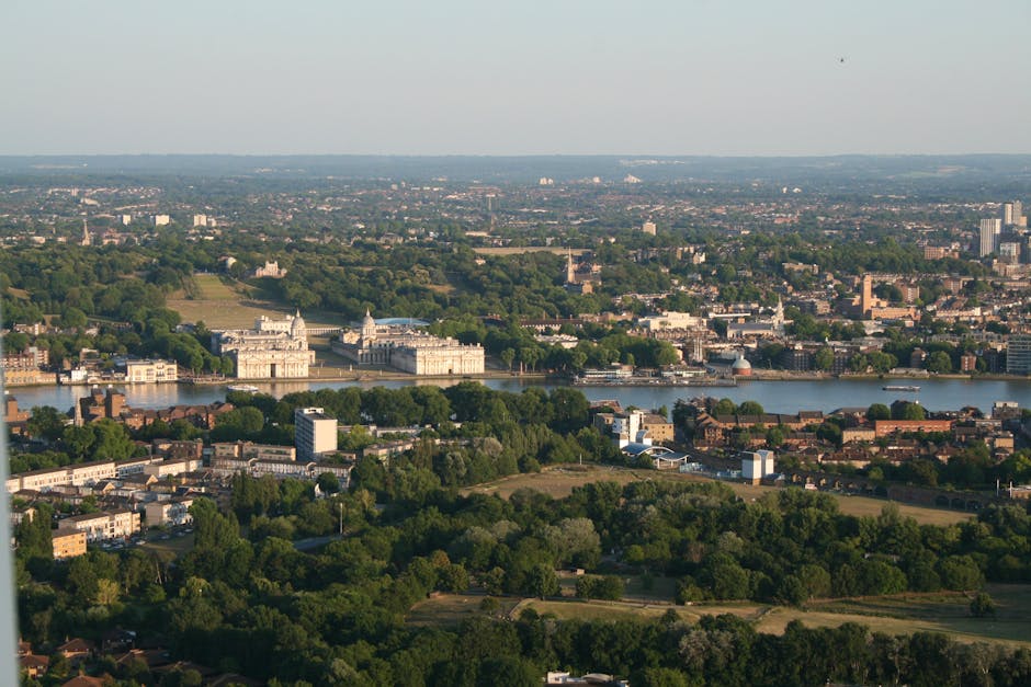 Aerial view of a cityscape showing a wide river flowing through the centre, with several historic and modern buildings along the riverbanks. In the foreground, there are lush green trees and open park
