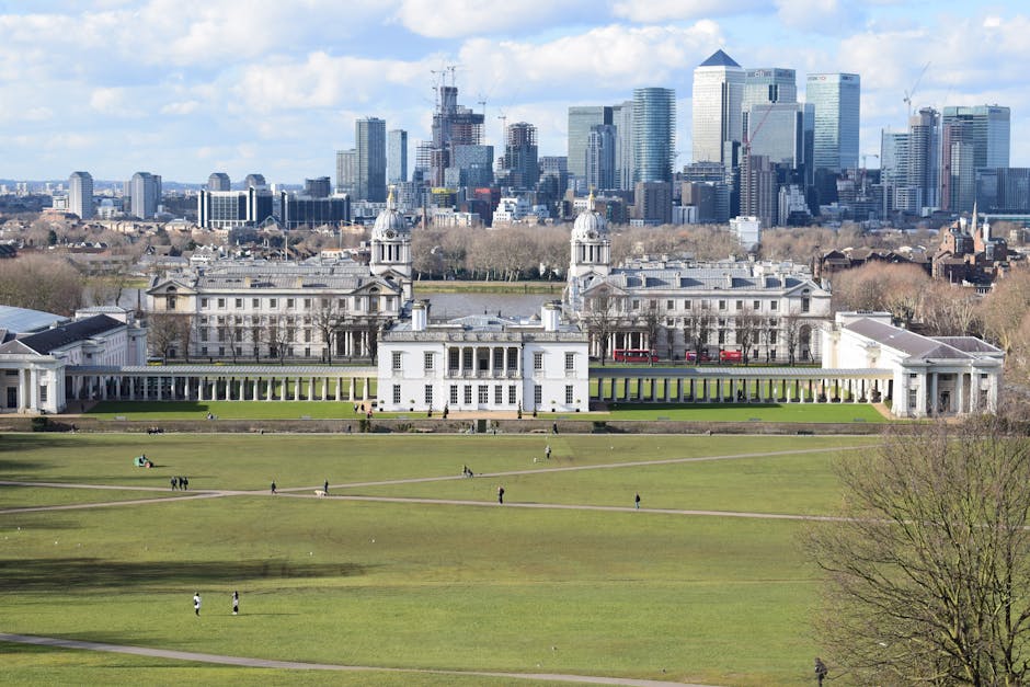 An aerial view of Greenwich with the River Thames prominently visible in the foreground, showing a mix of historic and modern buildings, including the Old Royal Naval College with its distinctive dome