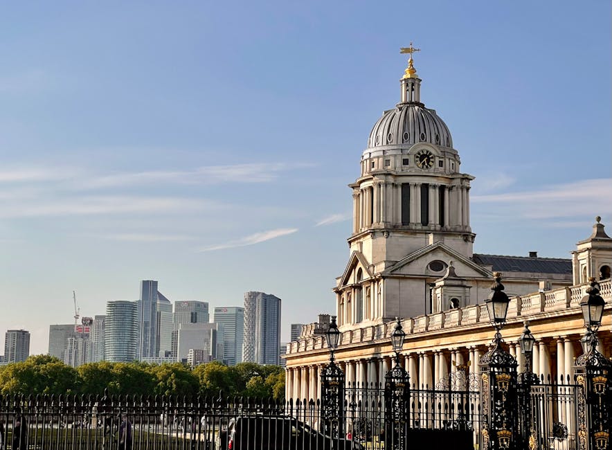 A wide view of the historic Old Royal Naval College in Greenwich, showing two symmetrical domed towers connected by a colonnaded terrace made of stone. The foreground features a paved pathway leading
