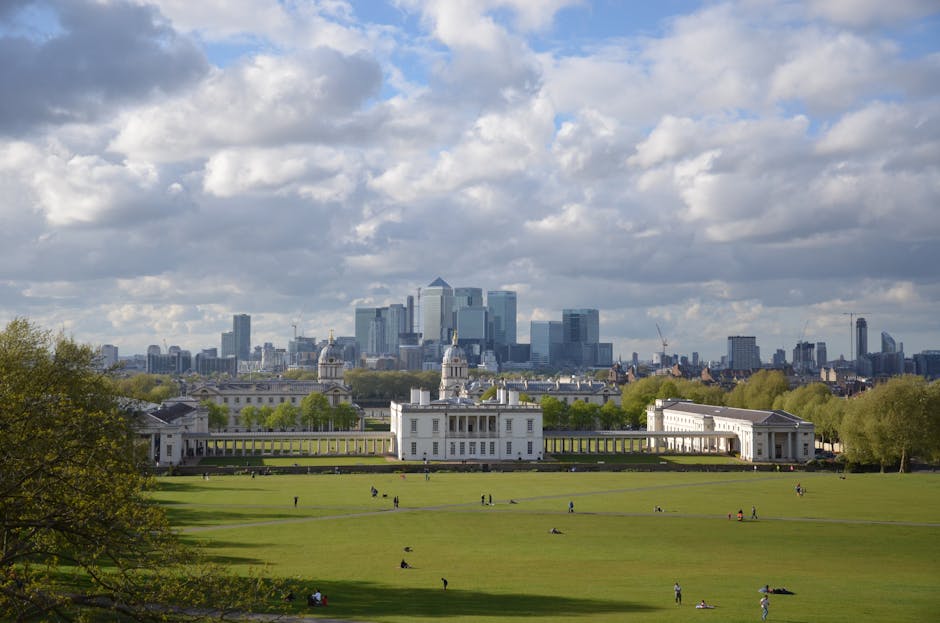 A city skyline with numerous modern high-rise buildings under construction, including cranes visible atop some towers, viewed from a park with lush green trees in the foreground. The scene captures an